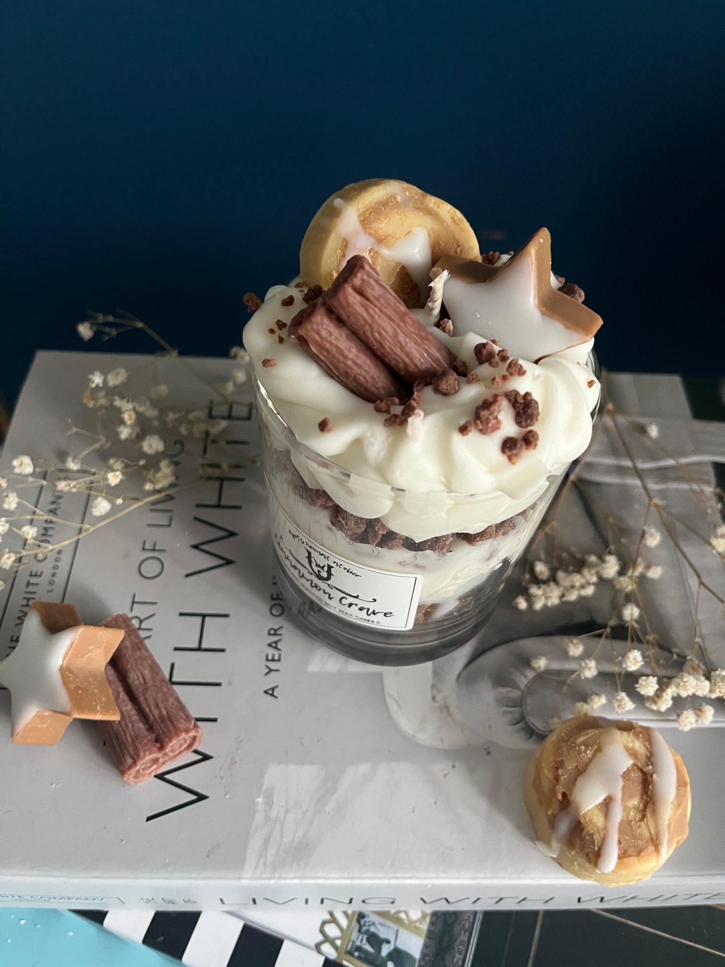 Eine dekorative Zimt Duftkerze in einem Glas, die wie ein Dessert gestaltet ist. Die Kerze zeigt abwechselnde Schichten aus Sahne und Keksboden, alles aus Wachs gefertigt. Obenauf befinden sich detailreiche Wachsdekorationen in Form einer Zimtschnecke, einer Zimtstange und eines Zimtsterns. Das Glas steht auf einem Stapel Bücher und ist von Trockenblumen umgeben, was eine gemütliche und herbstliche Atmosphäre schafft.