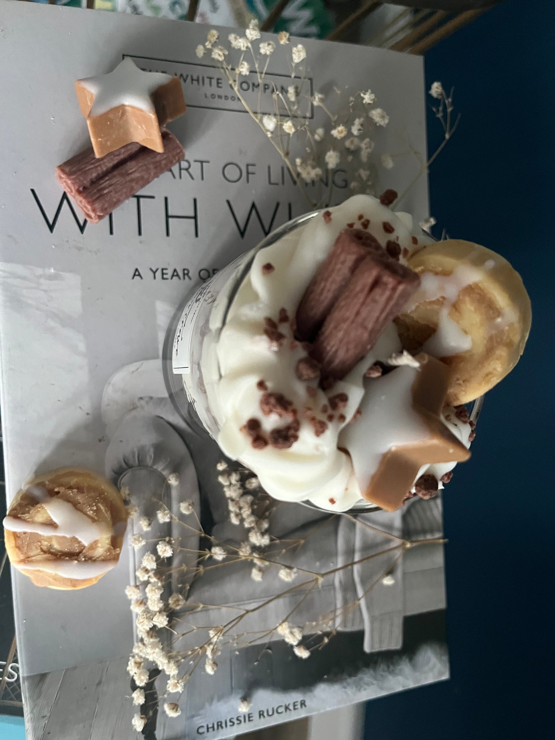 Eine dekorative Zimt-Duftkerze in einem Glas, die wie ein Dessert gestaltet ist. Die Kerze zeigt abwechselnde Schichten aus Sahne und Keksboden, alles aus Wachs gefertigt. Obenauf befinden sich detailreiche Wachsdekorationen in Form einer Zimtschnecke, einer Zimtstange und eines Zimtsterns. Das Glas steht auf einem Stapel Bücher und ist von Trockenblumen umgeben, was eine gemütliche und herbstliche Atmosphäre schafft.