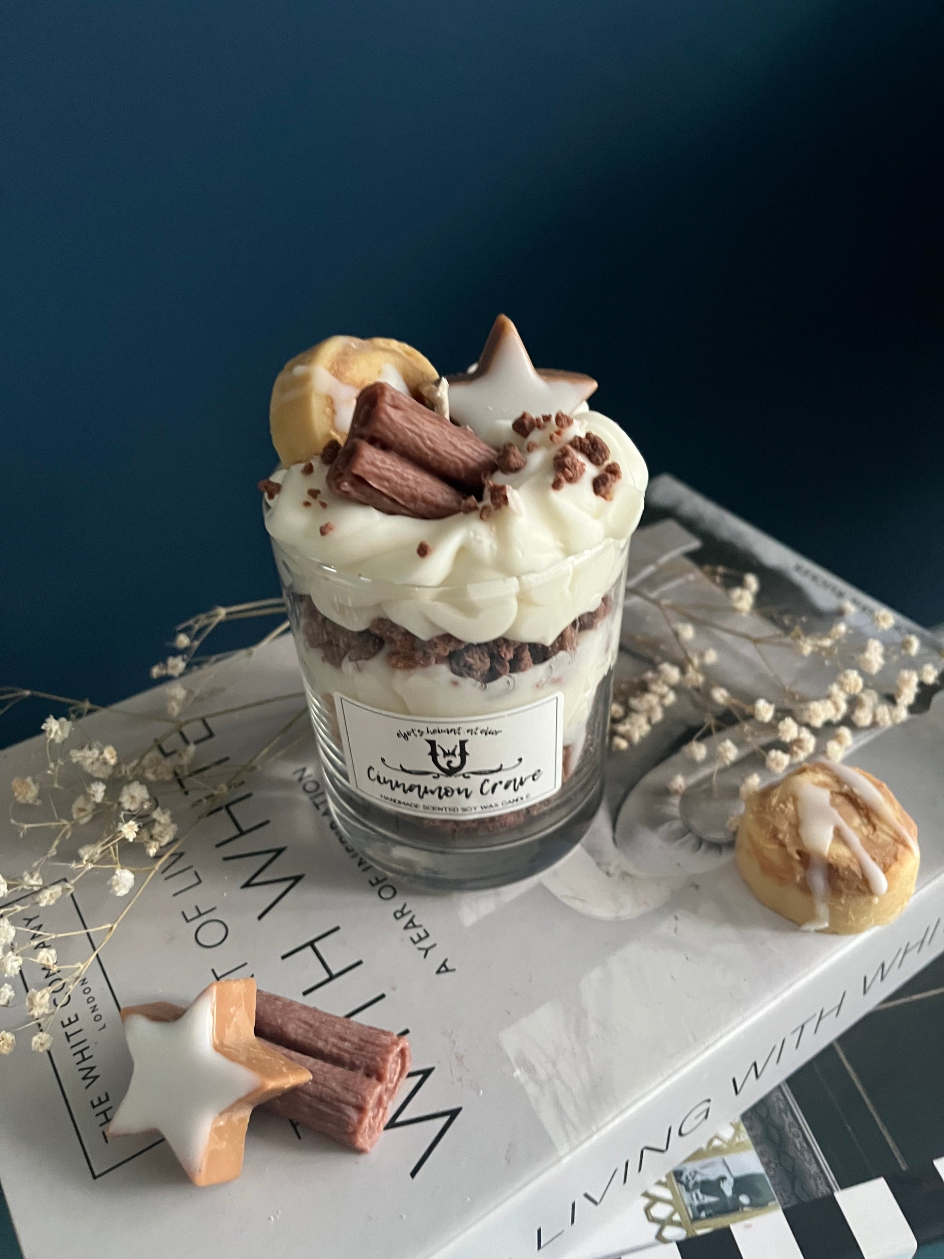 Eine dekorative Zimt-Duftkerze in einem Glas, die wie ein Dessert gestaltet ist. Die Kerze zeigt abwechselnde Schichten aus Sahne und Keksboden, alles aus Wachs gefertigt. Obenauf befinden sich detailreiche Wachsdekorationen in Form einer Zimtschnecke, einer Zimtstange und eines Zimtsterns. Das Glas steht auf einem Stapel Bücher und ist von Trockenblumen umgeben, was eine gemütliche und herbstliche Atmosphäre schafft.